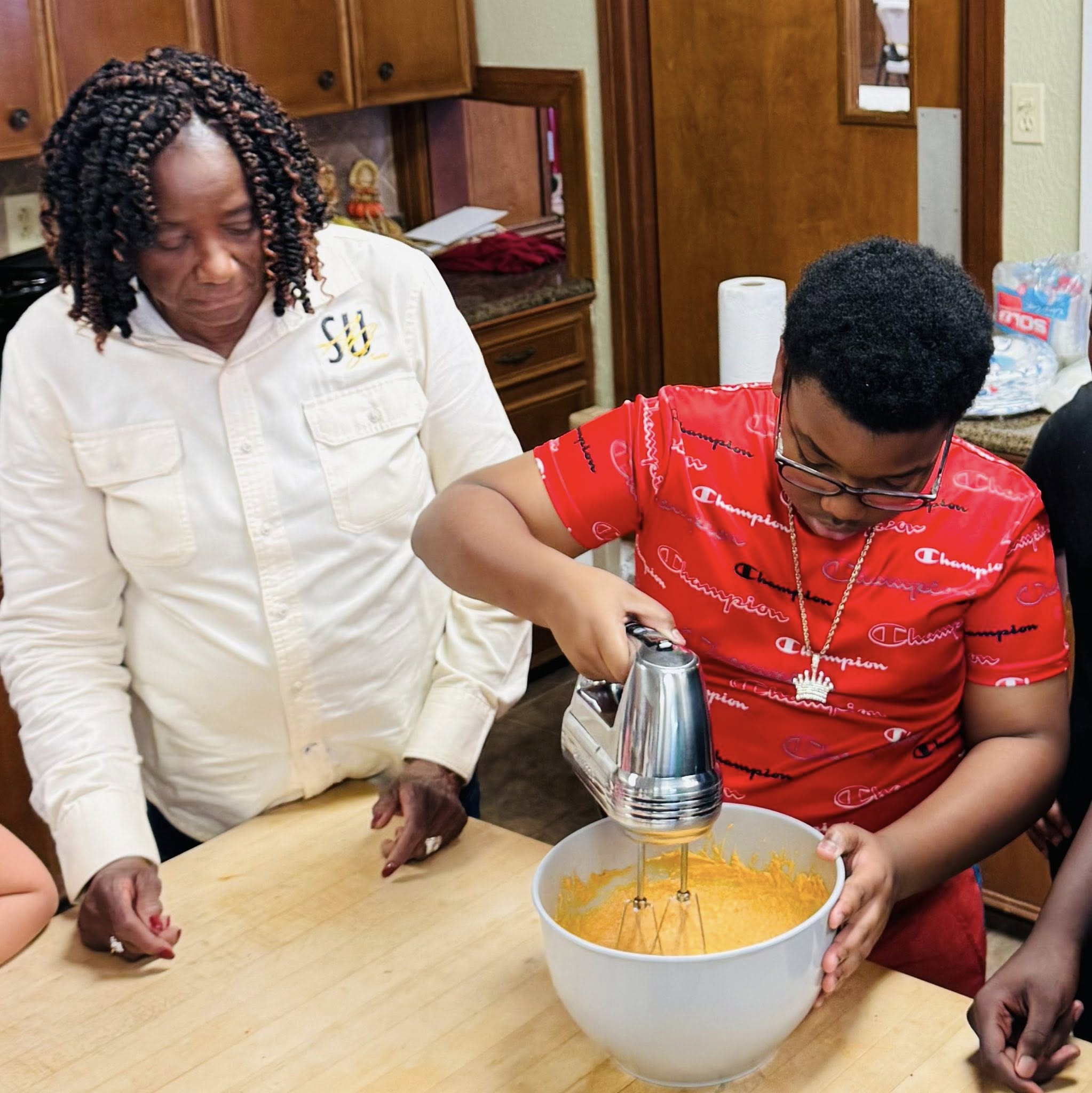 A woman watching a boy use a mixer in a bowl.